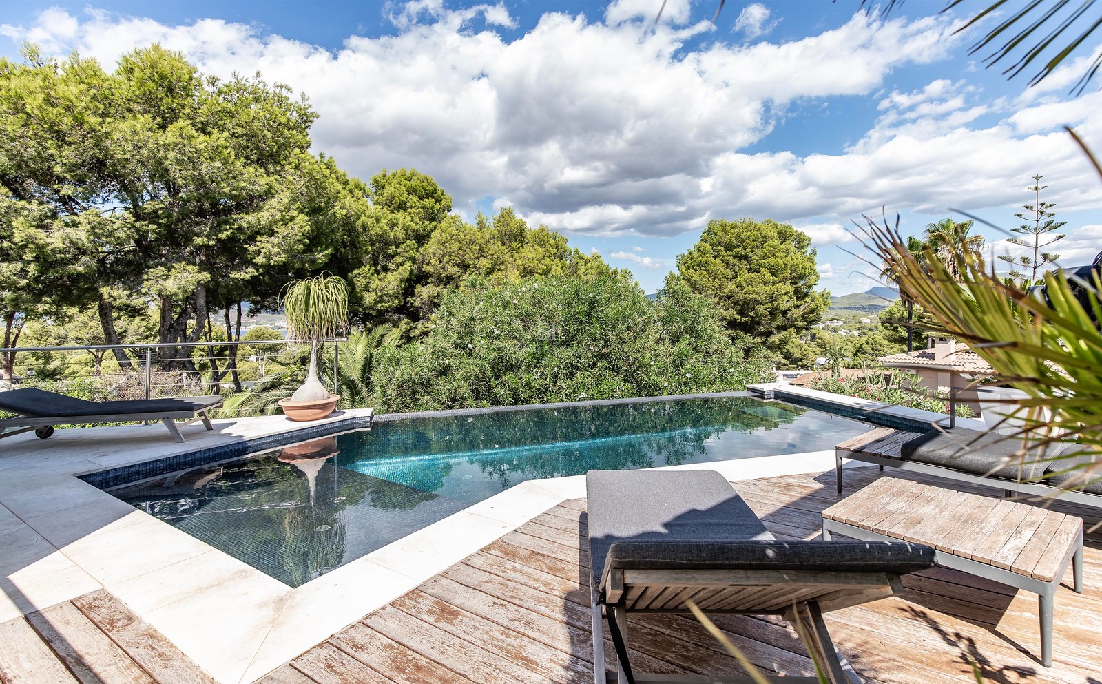 Une piscine à débordement aux carreaux sombres entourée d'une terrasse en bois et de bains de soleil.
