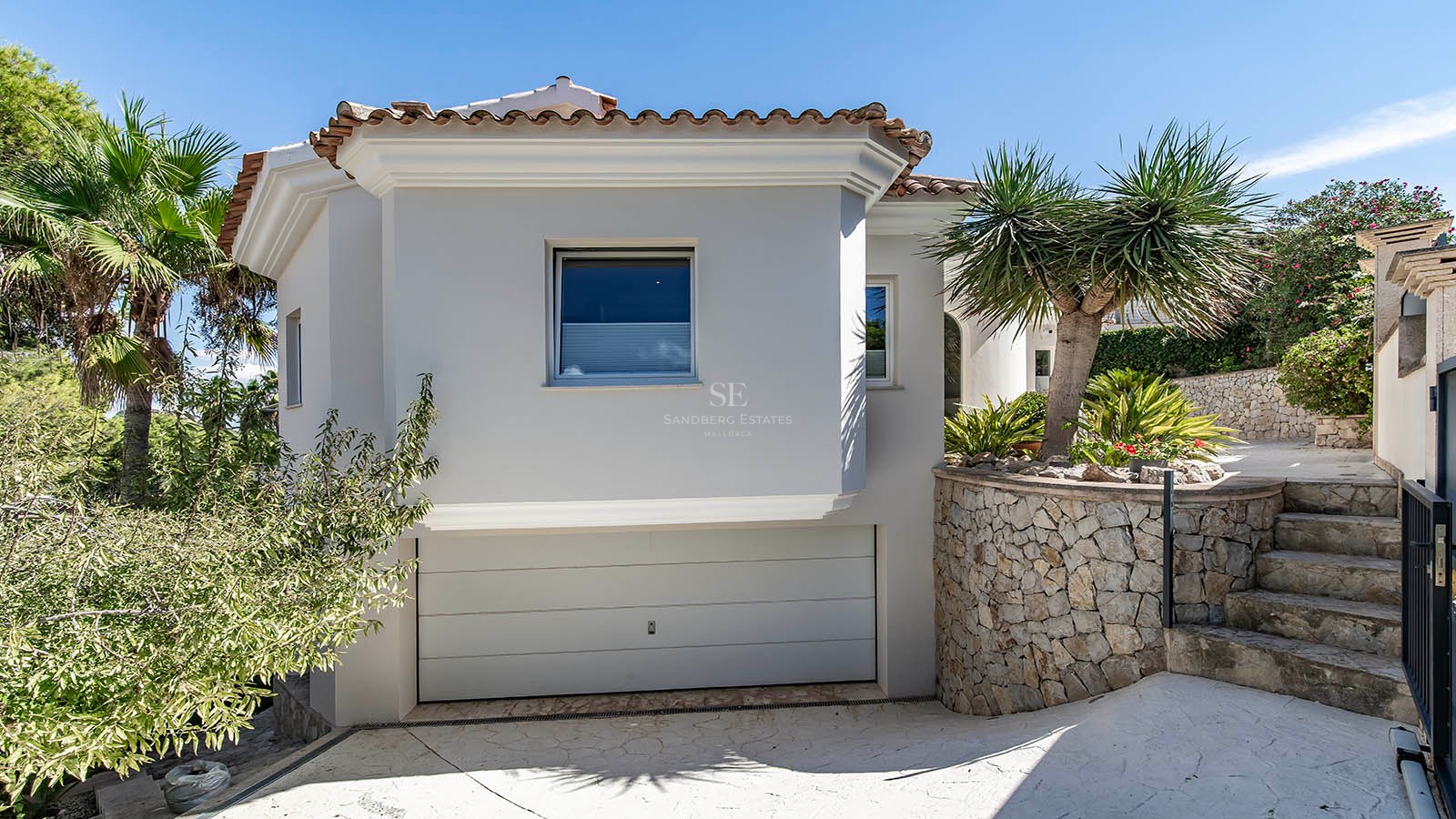 Extérieur de villa méditerranéenne blanche avec mur en pierre naturelle, garage intégré et aménagement paysager tropical.