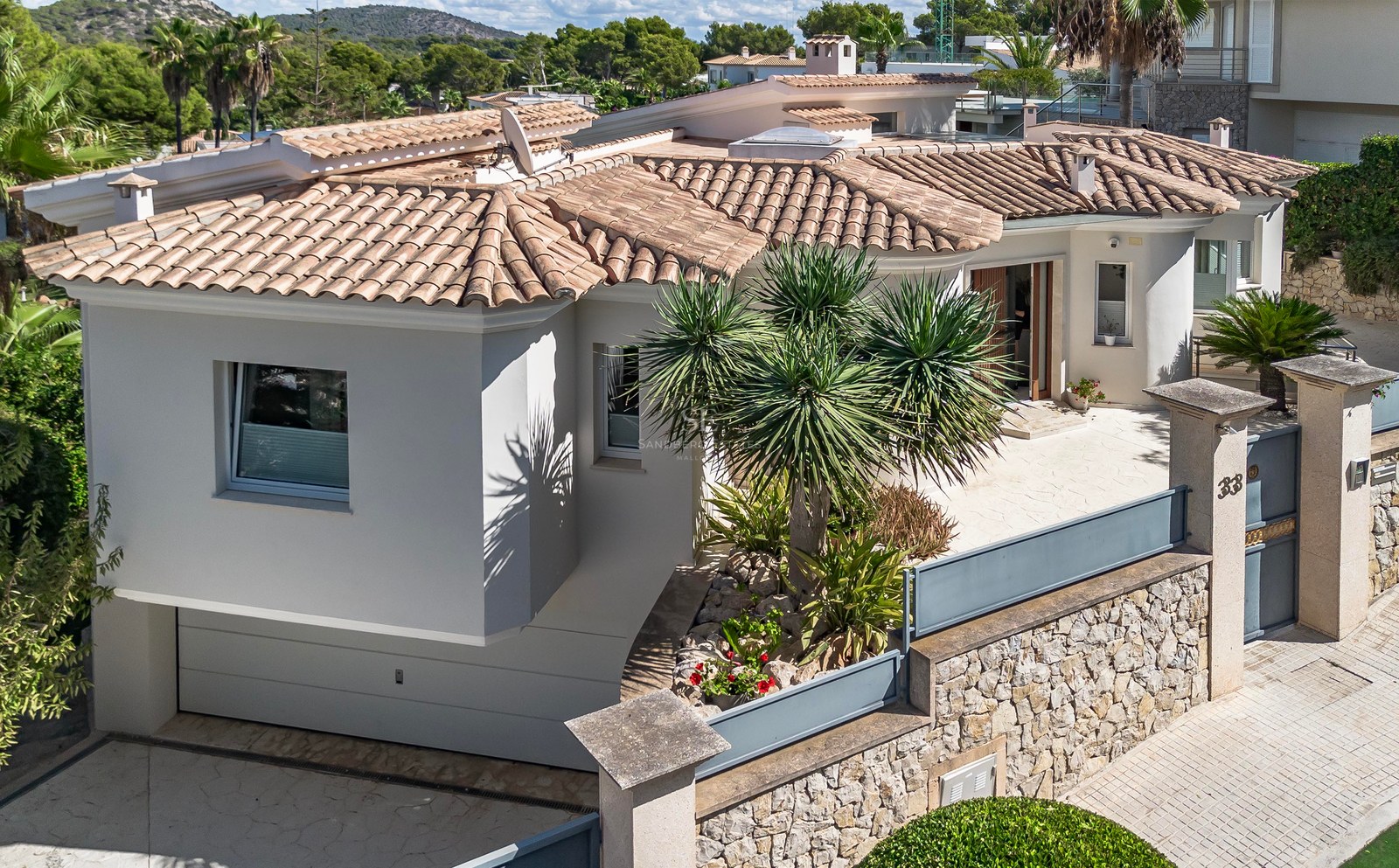 Vue en plongée d'une villa blanche avec toit en tuiles, murs en pierre et jardin tropical sous le soleil.