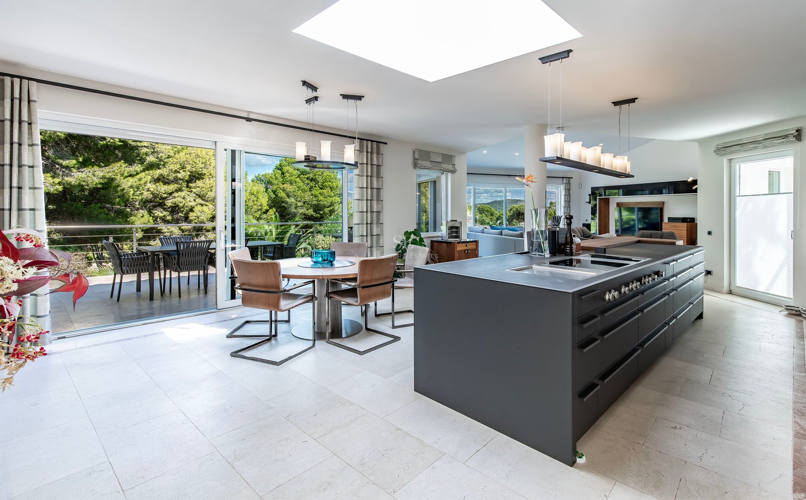 Cuisine moderne avec îlot noir mat, coin repas et grandes baies vitrées ouvrant sur une terrasse boisée.