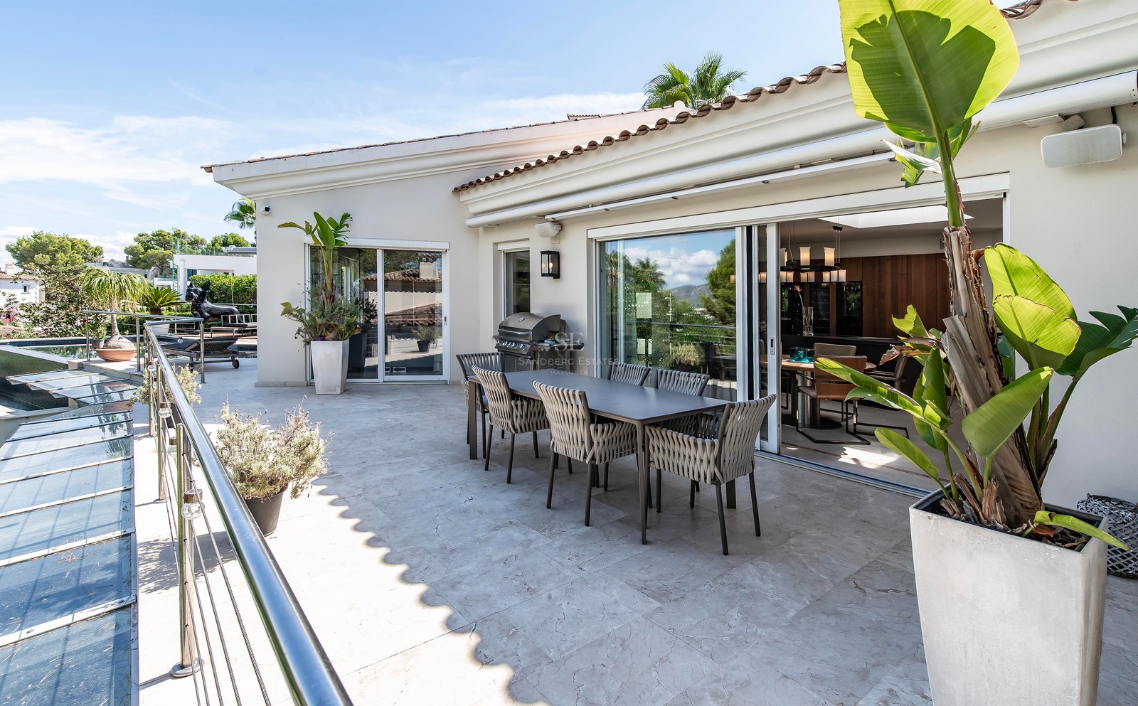 Terrasse ensoleillée avec dallage en pierre, table à manger et barbecue, s'ouvrant sur l'intérieur par des baies vitrées.