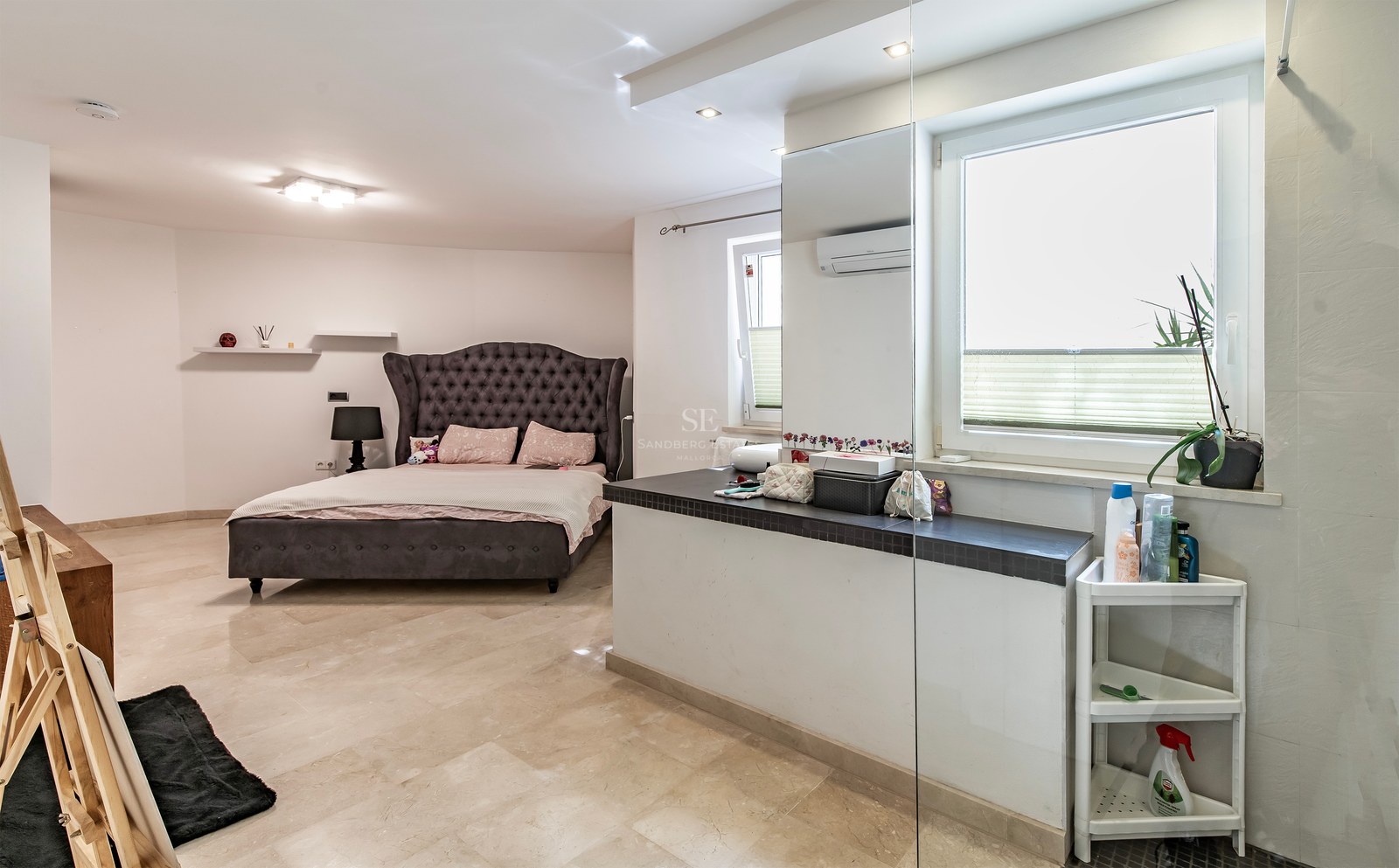 Grande chambre avec lit capitonné gris anthracite, sol en pierre crème et cloison avec rangement.