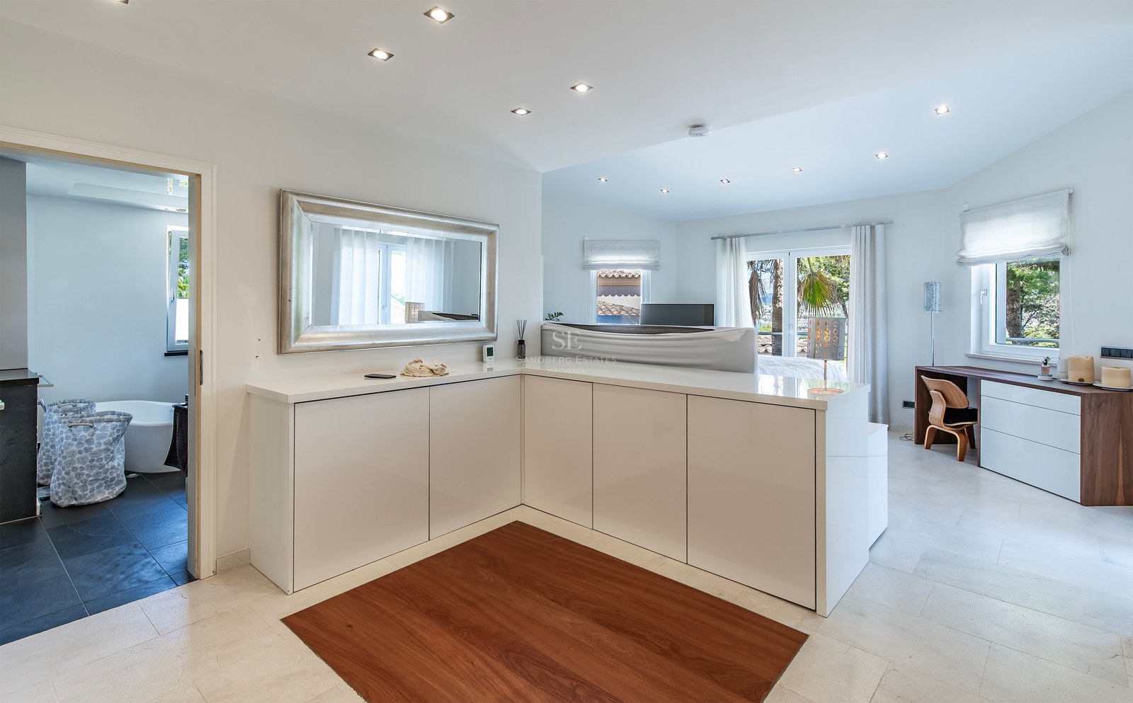 Grande chambre moderne avec rangements blancs, grand miroir, sol en pierre et vue sur la salle de bains attenante.