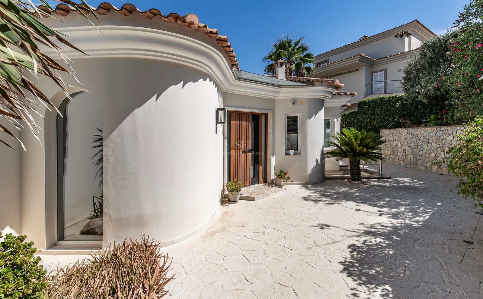 Extérieur de villa blanche aux lignes courbes, porte en bois, toit en terre cuite et allée pavée sous ciel bleu.