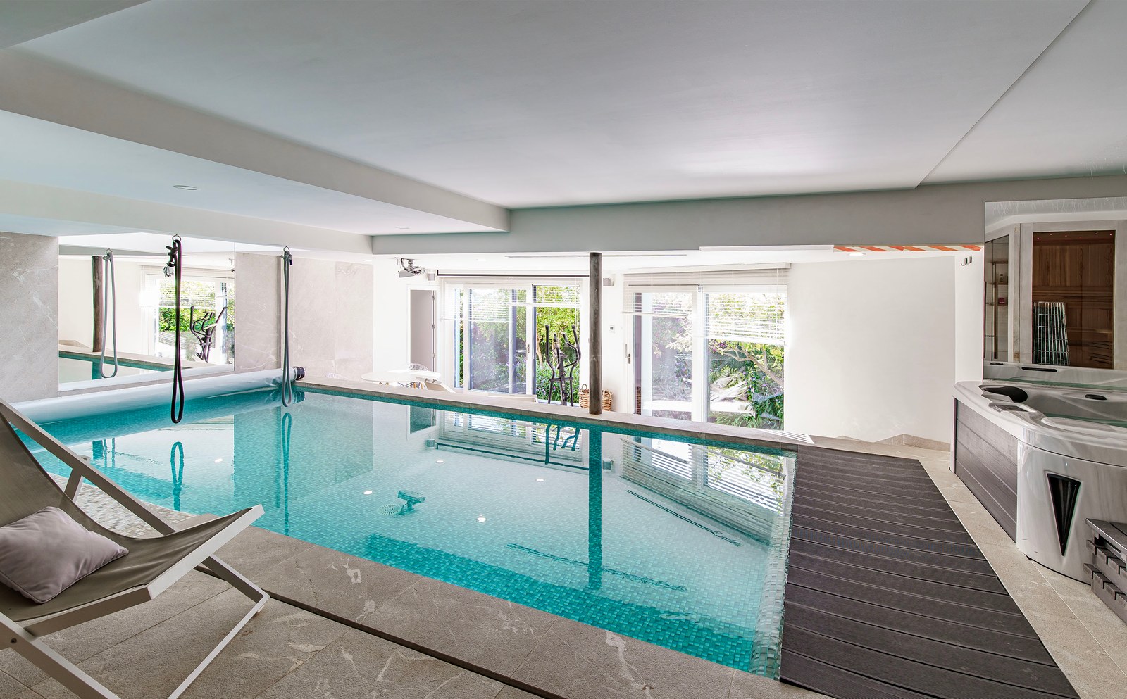 Piscina interior con mosaicos turquesa, jacuzzi y puertas de cristal hacia el jardín.