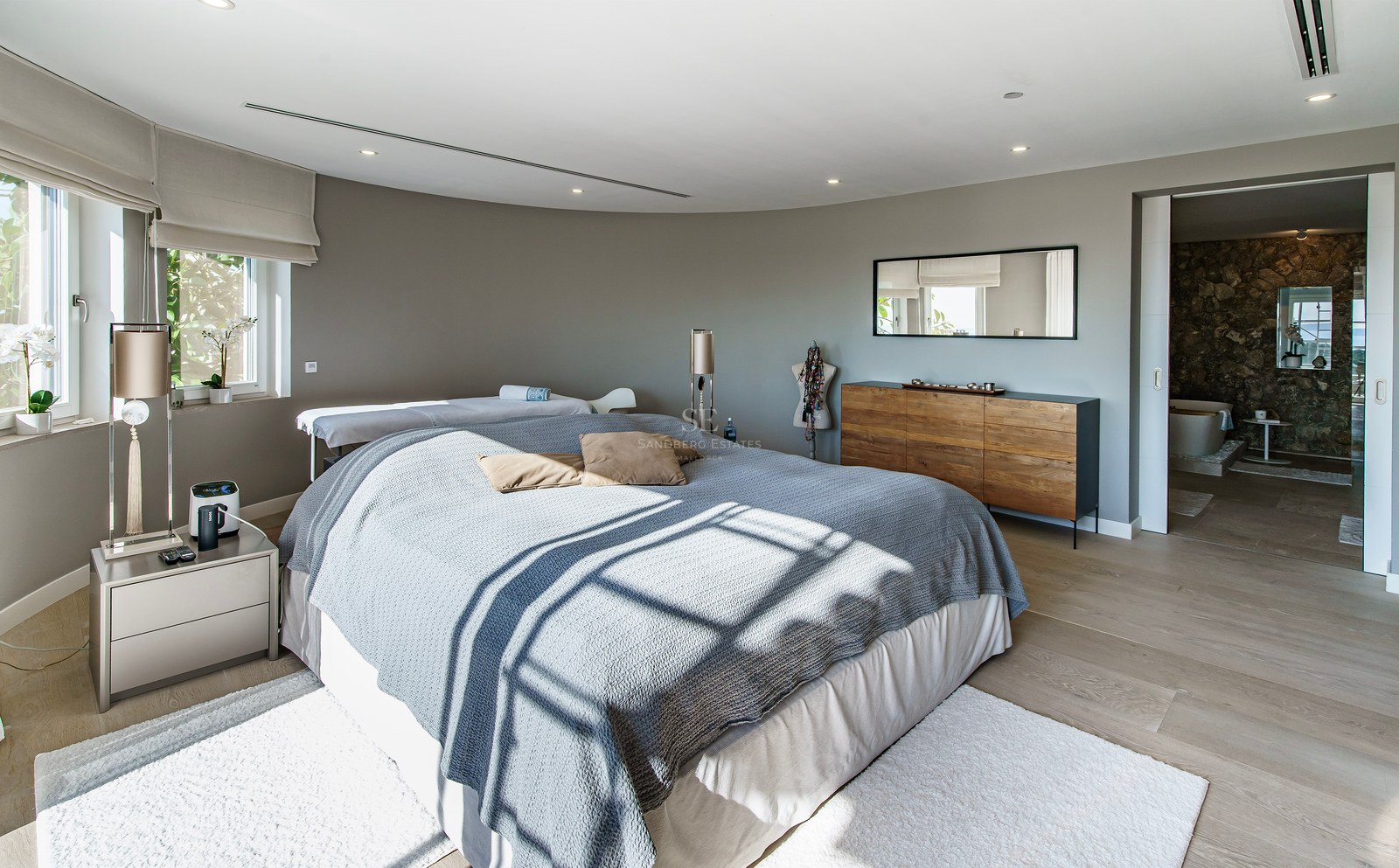 Dormitorio principal contemporáneo con ropa de cama gris, aparador de madera y vistas al baño privado con pared de piedra.