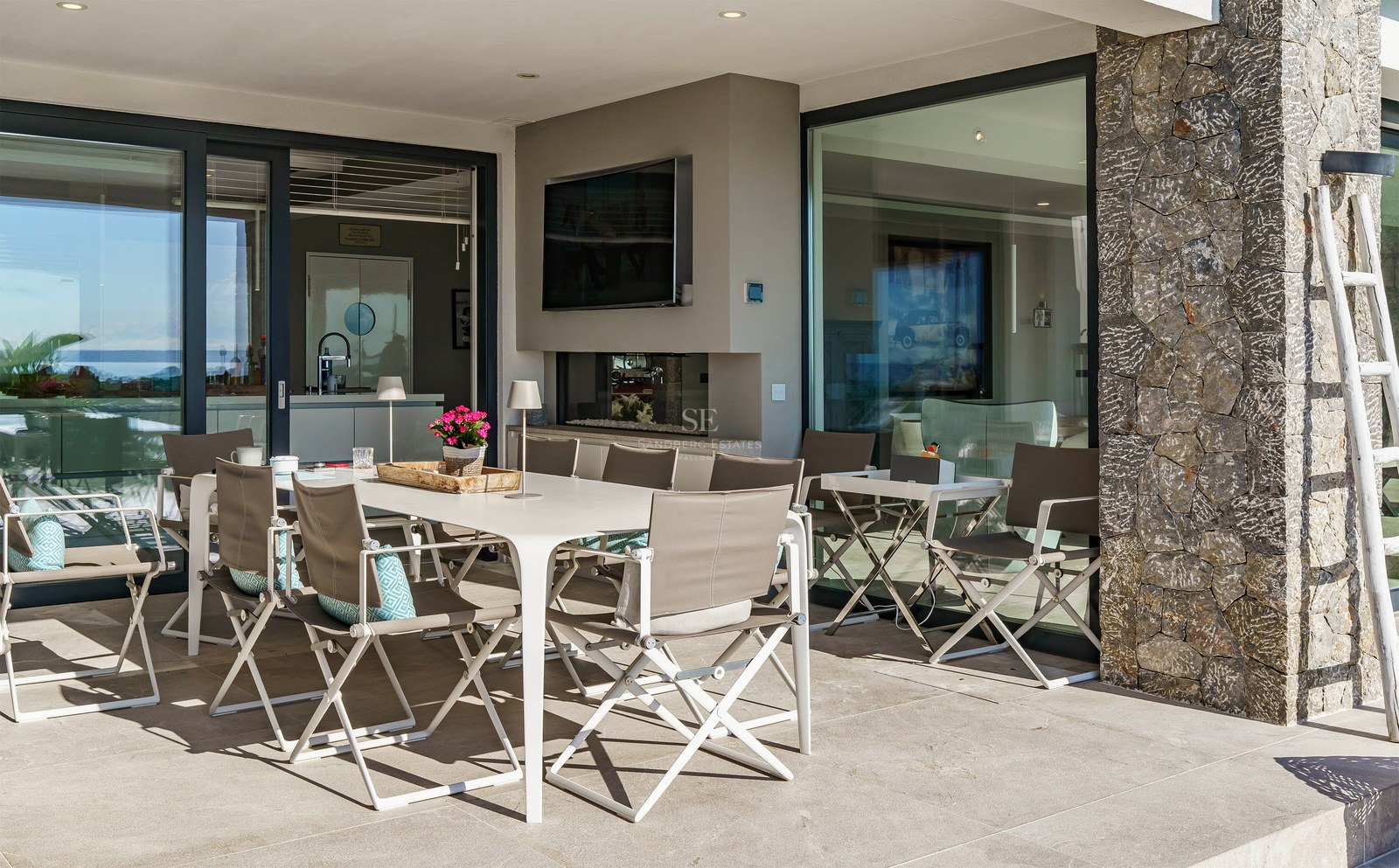 Terraza cubierta moderna con mesa blanca, sillas grises, pared de piedra natural y puertas de cristal con vistas al mar.