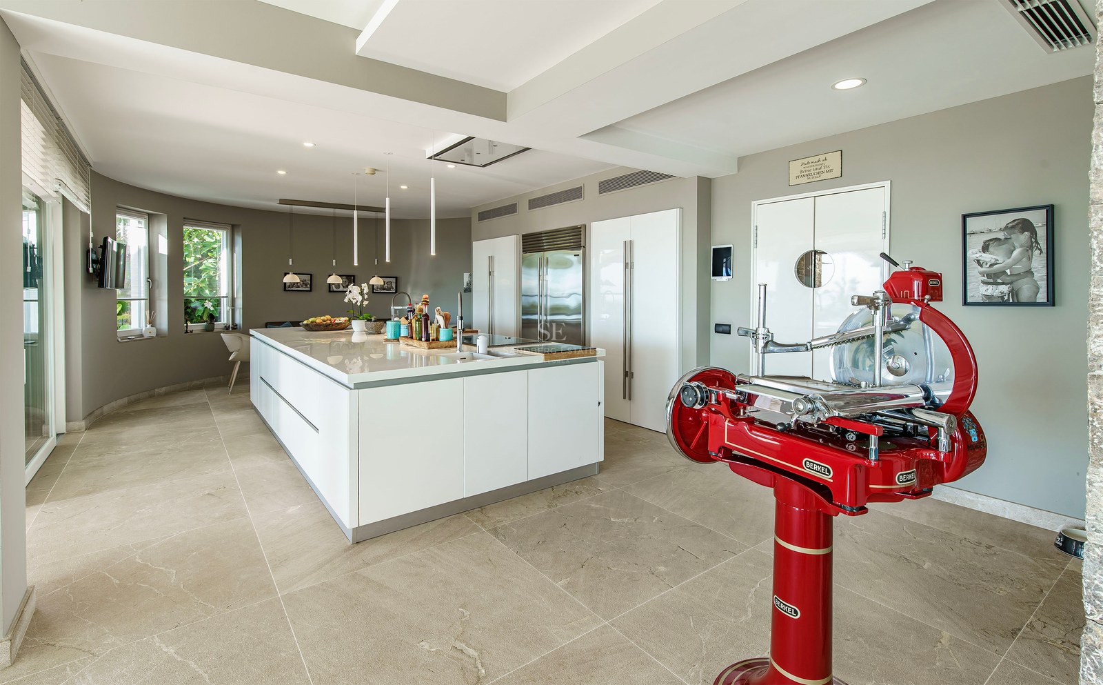Cocina moderna blanca con isla grande, suelo de piedra, electrodomésticos integrados y cortadora de fiambre roja.
