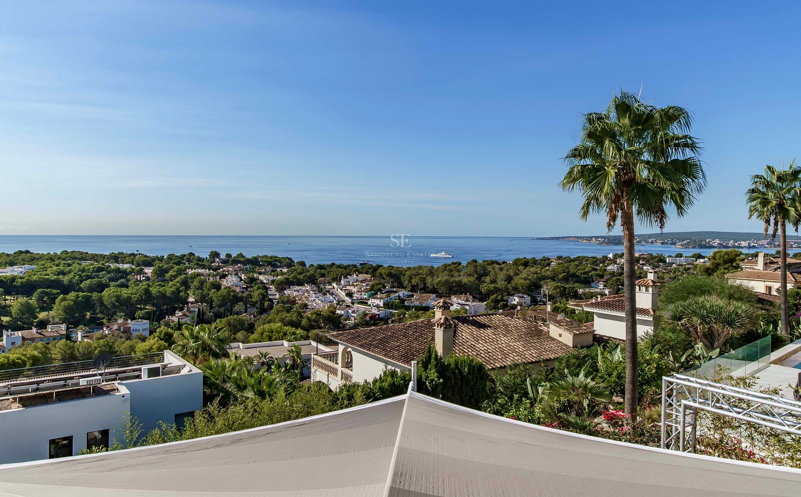 Vista elevada del mar azul y villas costeras rodeadas de vegetación y palmeras bajo un cielo despejado.