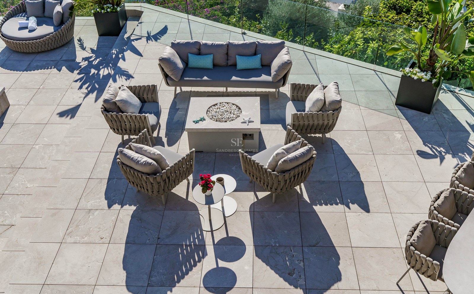 Vista aérea de una terraza de piedra con muebles de diseño, un brasero central y barandillas de cristal.