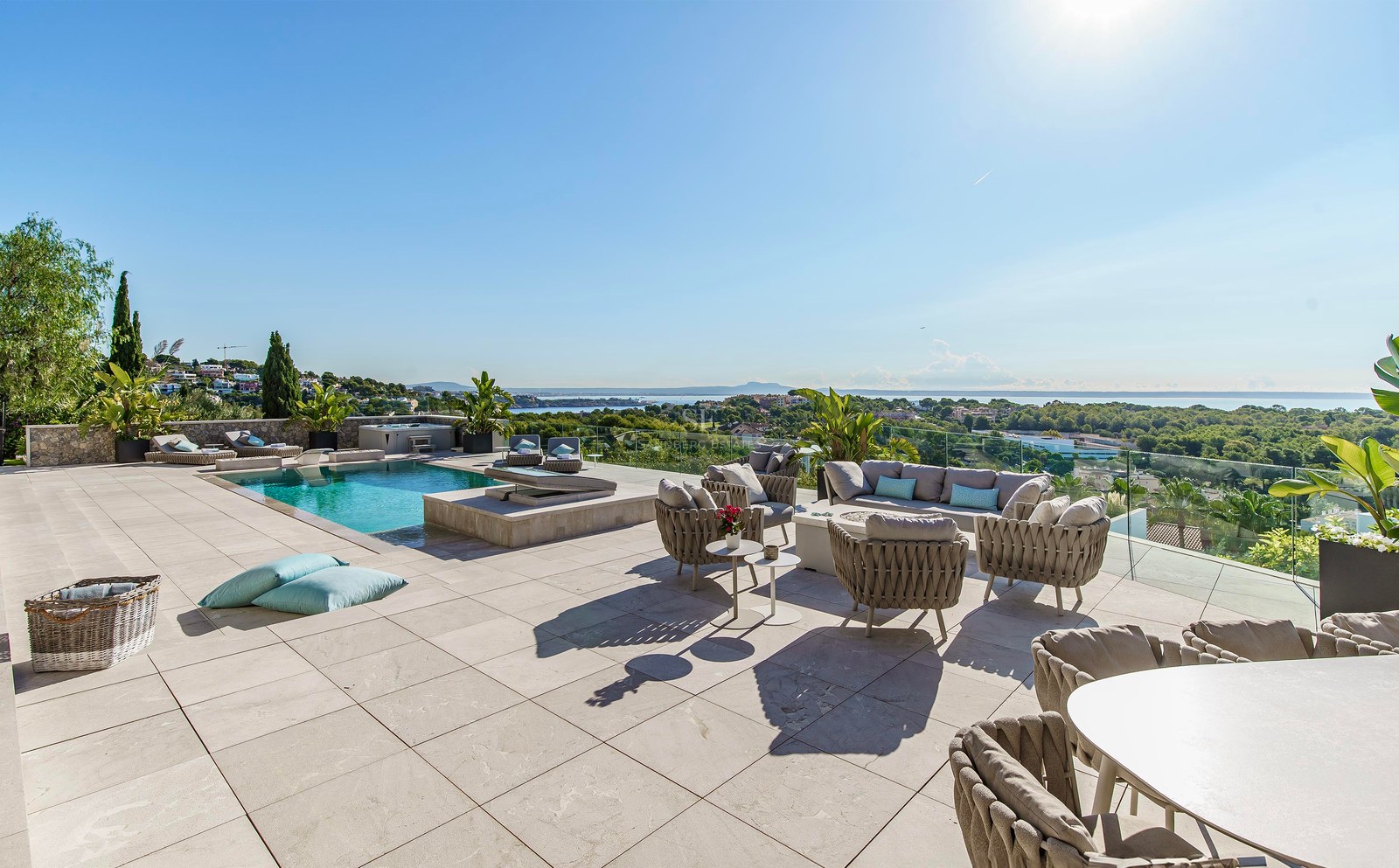 Terraza de piedra con muebles de diseño, piscina y vistas panorámicas al mar y al paisaje verde bajo un cielo despejado.