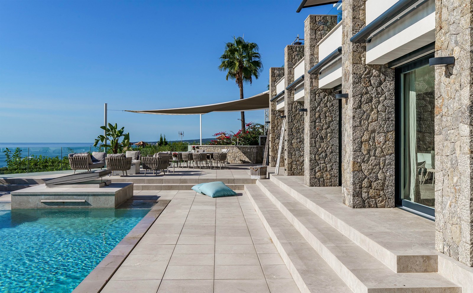 Terraza de piedra con piscina infinity, zona de estar sombreada y vistas panorámicas al mar bajo un cielo despejado.