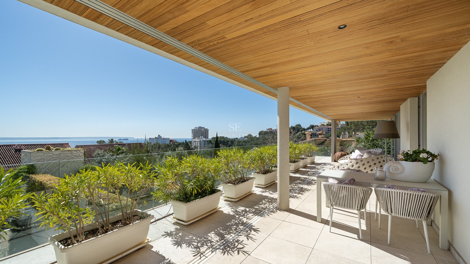 A spacious covered terrace with a wooden slatted ceiling, white planters, and a dining area overlooking the sea.