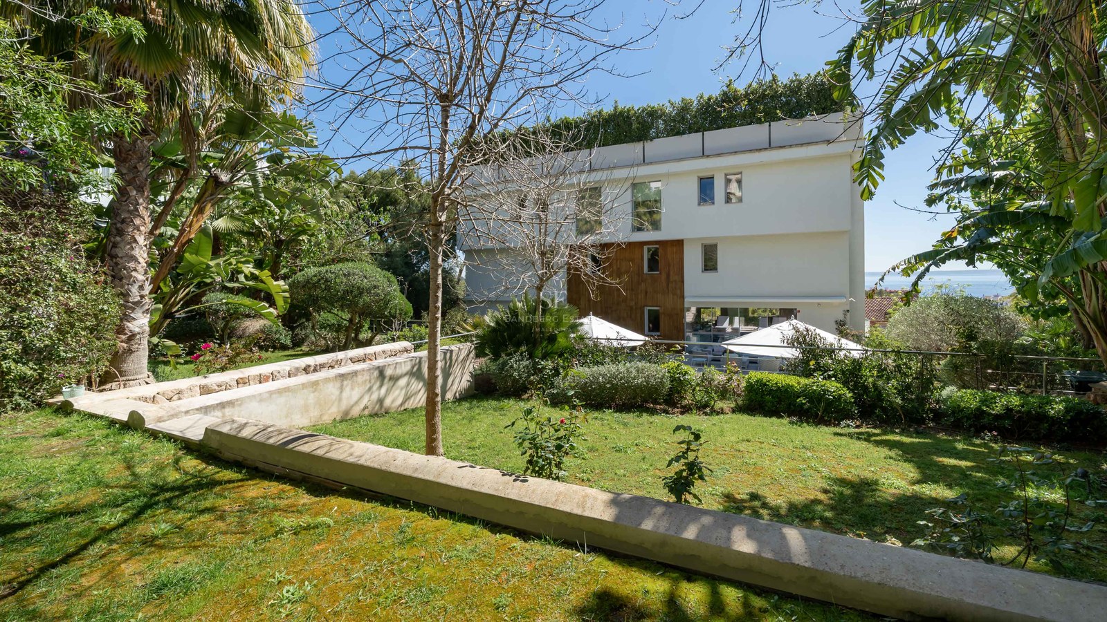 Modern white three-story villa with wood paneling accents viewed from a manicured green garden with stone walls.