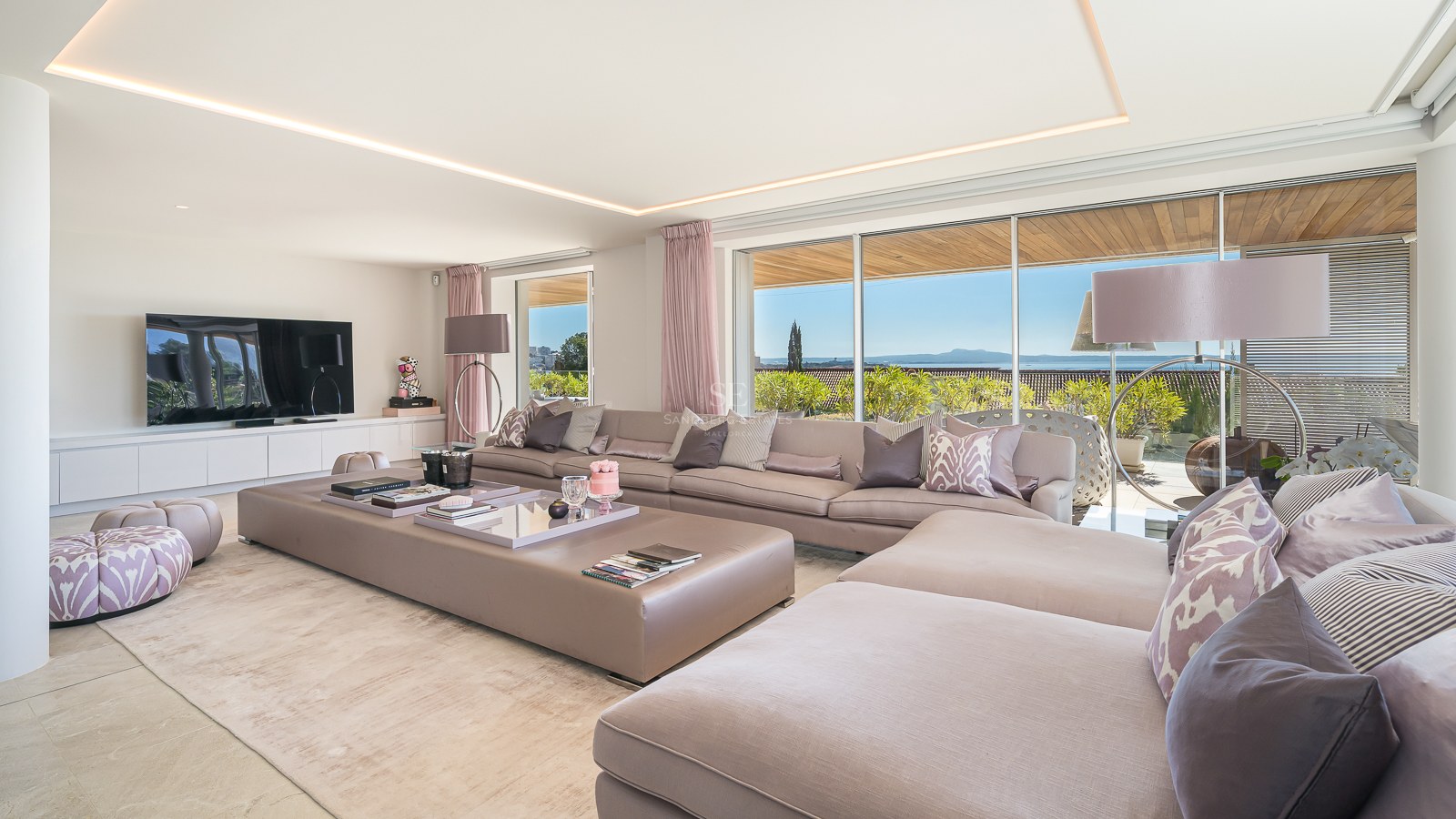 Spacious modern living room with large modular taupe sofas, natural stone flooring, and floor-to-ceiling windows with sea views.