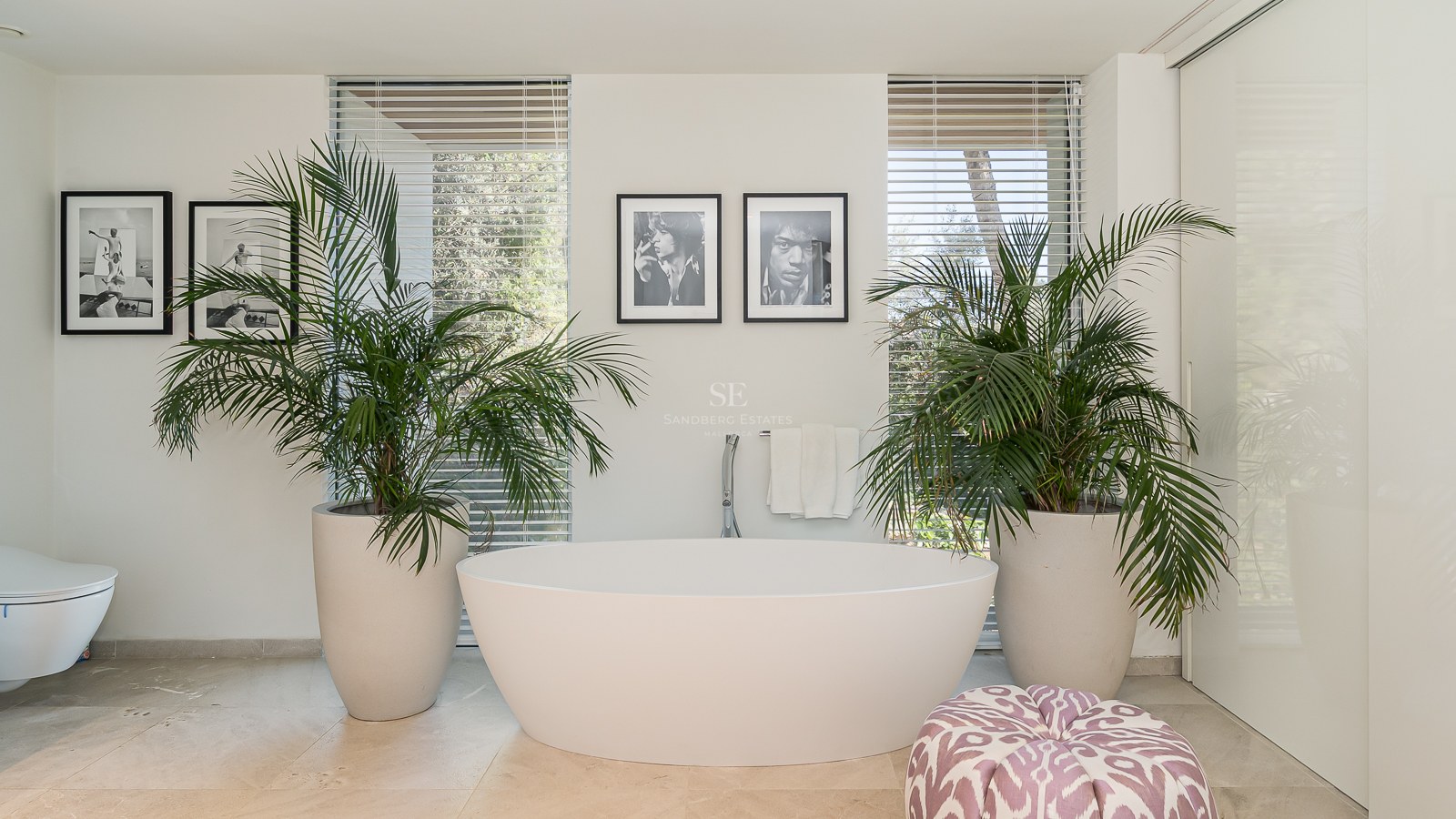 Modern master bathroom featuring a white freestanding oval bathtub flanked by large potted palms and natural light.