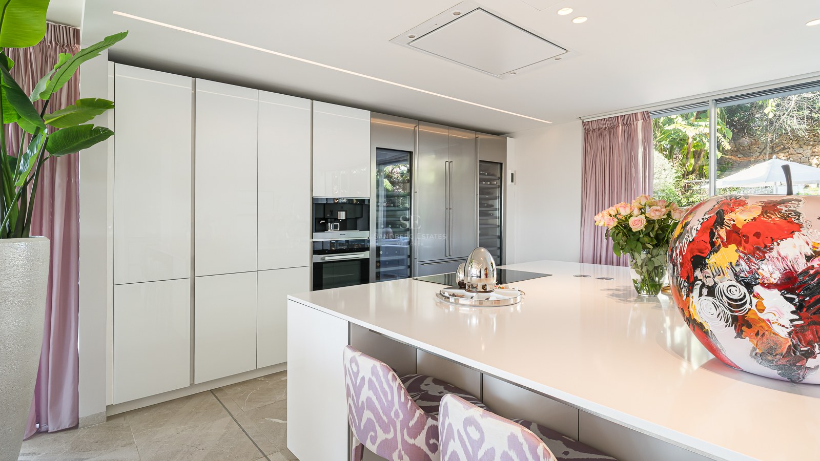 Bright modern kitchen featuring a white central island, floor-to-ceiling cabinetry, and large windows with garden views.