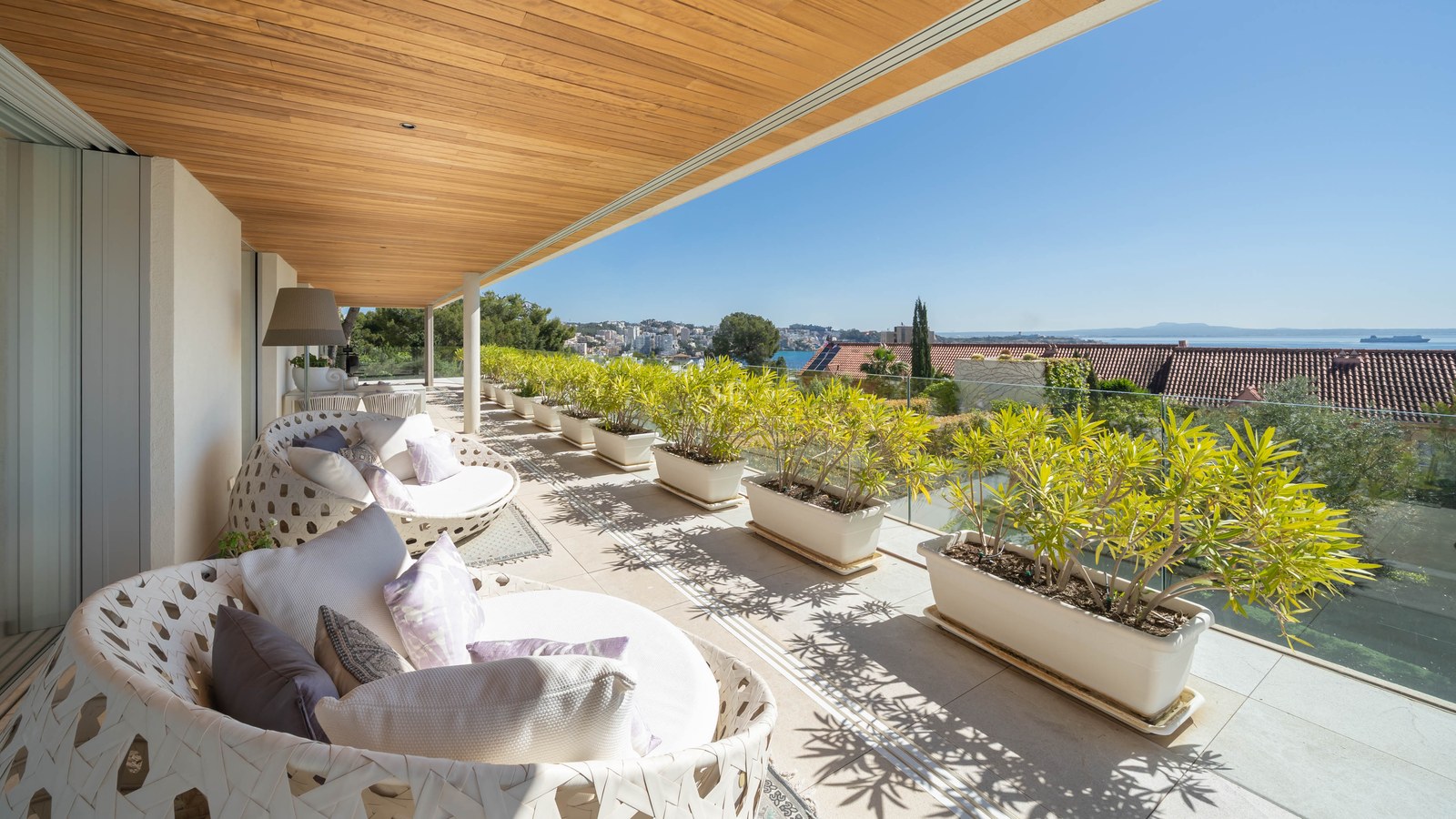 Modern covered terrace with wooden ceiling, white lounge furniture, and potted plants overlooking the sea.