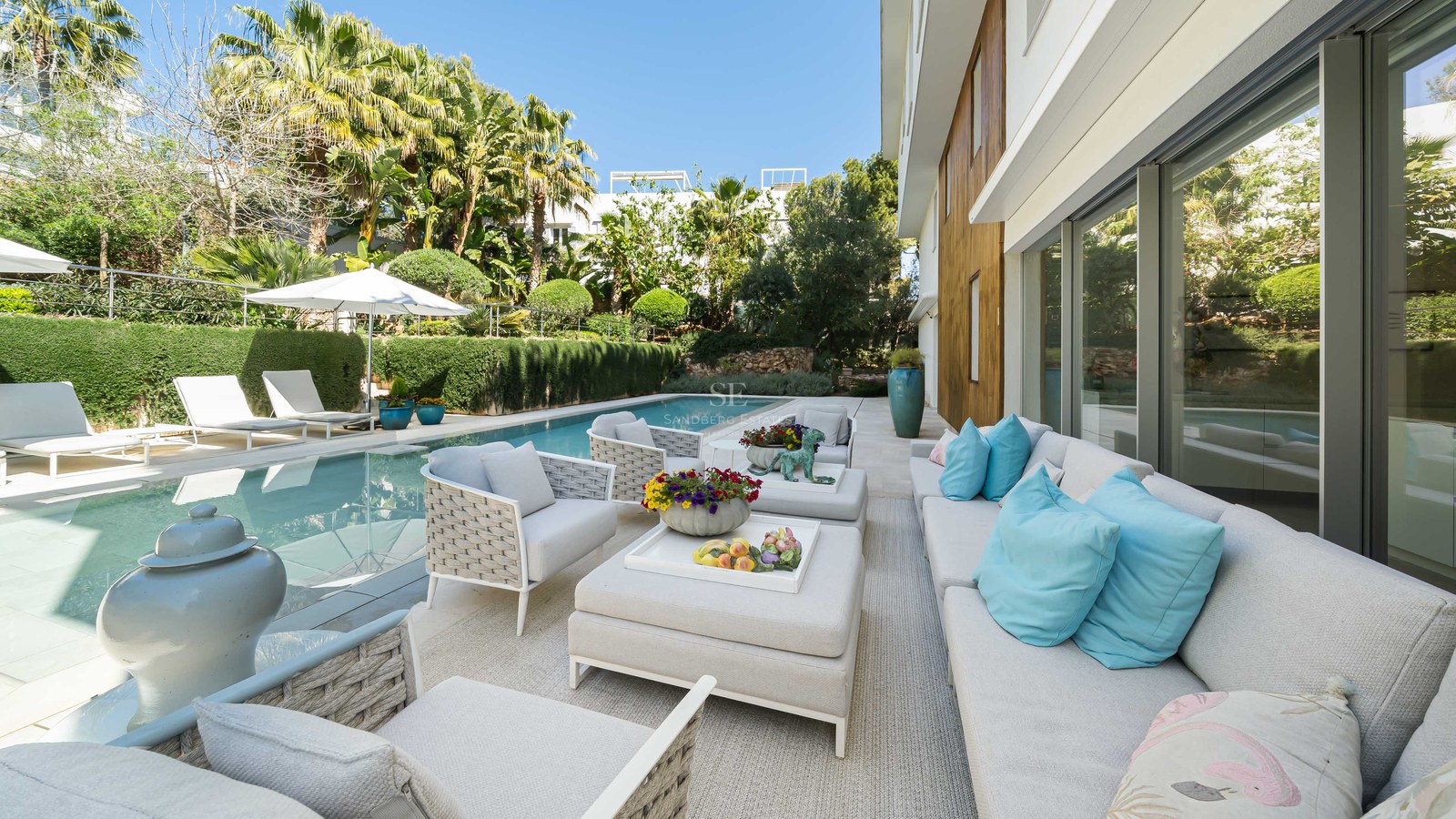 Luxury rectangular swimming pool alongside a stone terrace with white designer sofas, turquoise cushions, and lush palms.