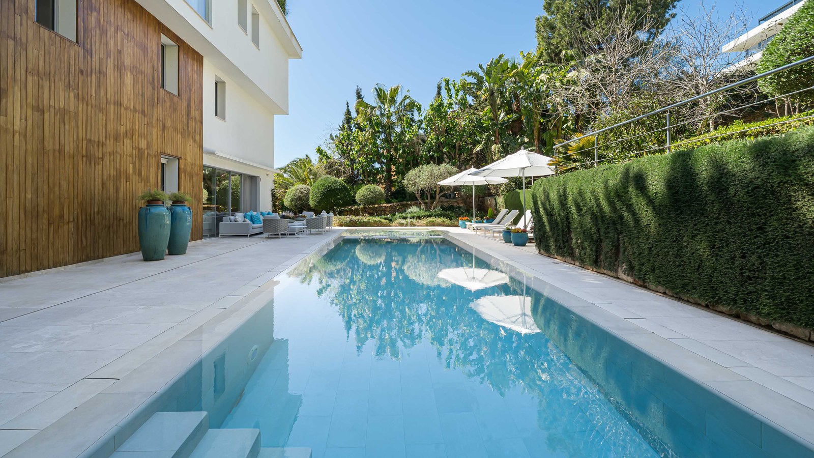 A long turquoise swimming pool surrounded by a cream stone terrace, wood-paneled walls, and vibrant green hedges.