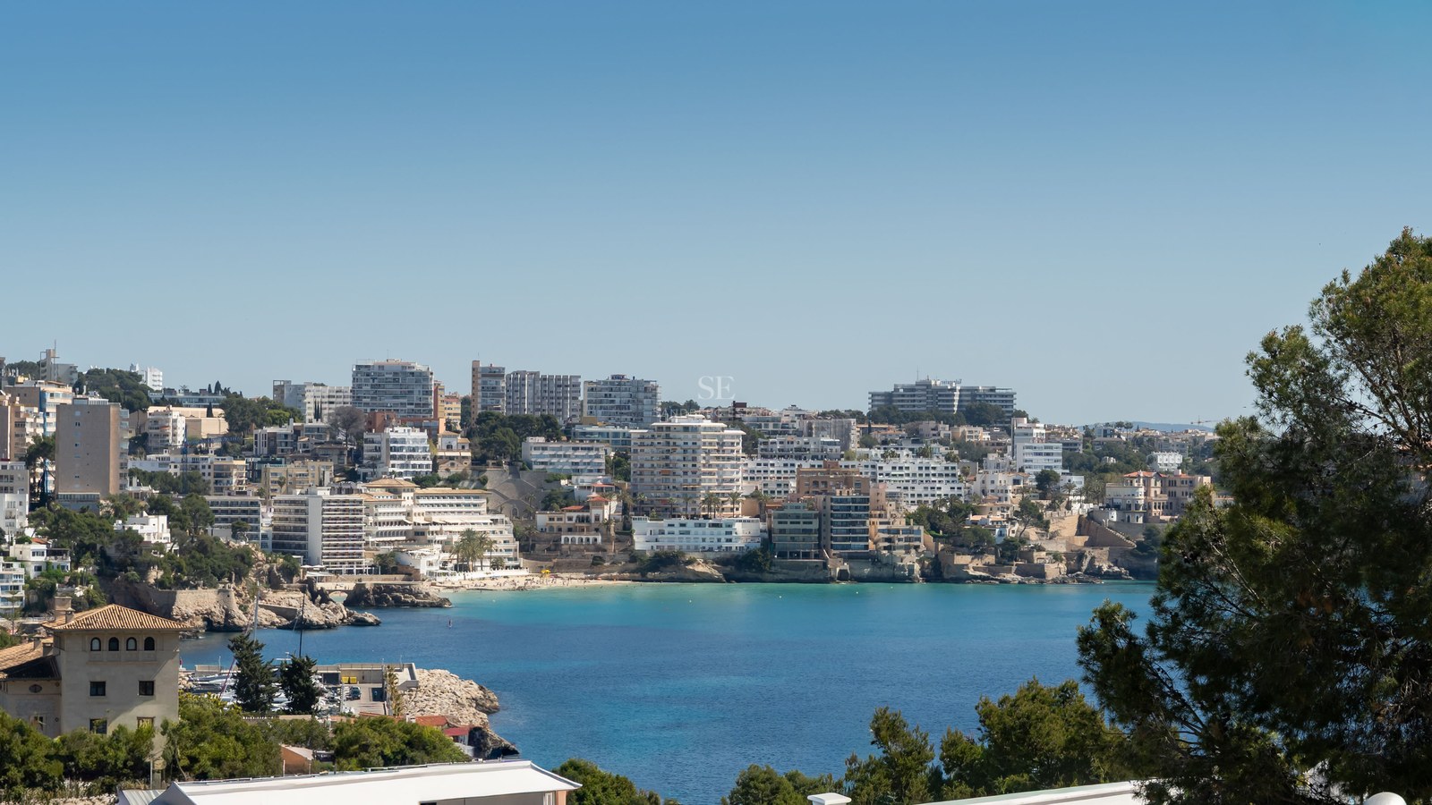 Elevated view of a turquoise bay surrounded by white coastal architecture and lush greenery under a clear blue sky.