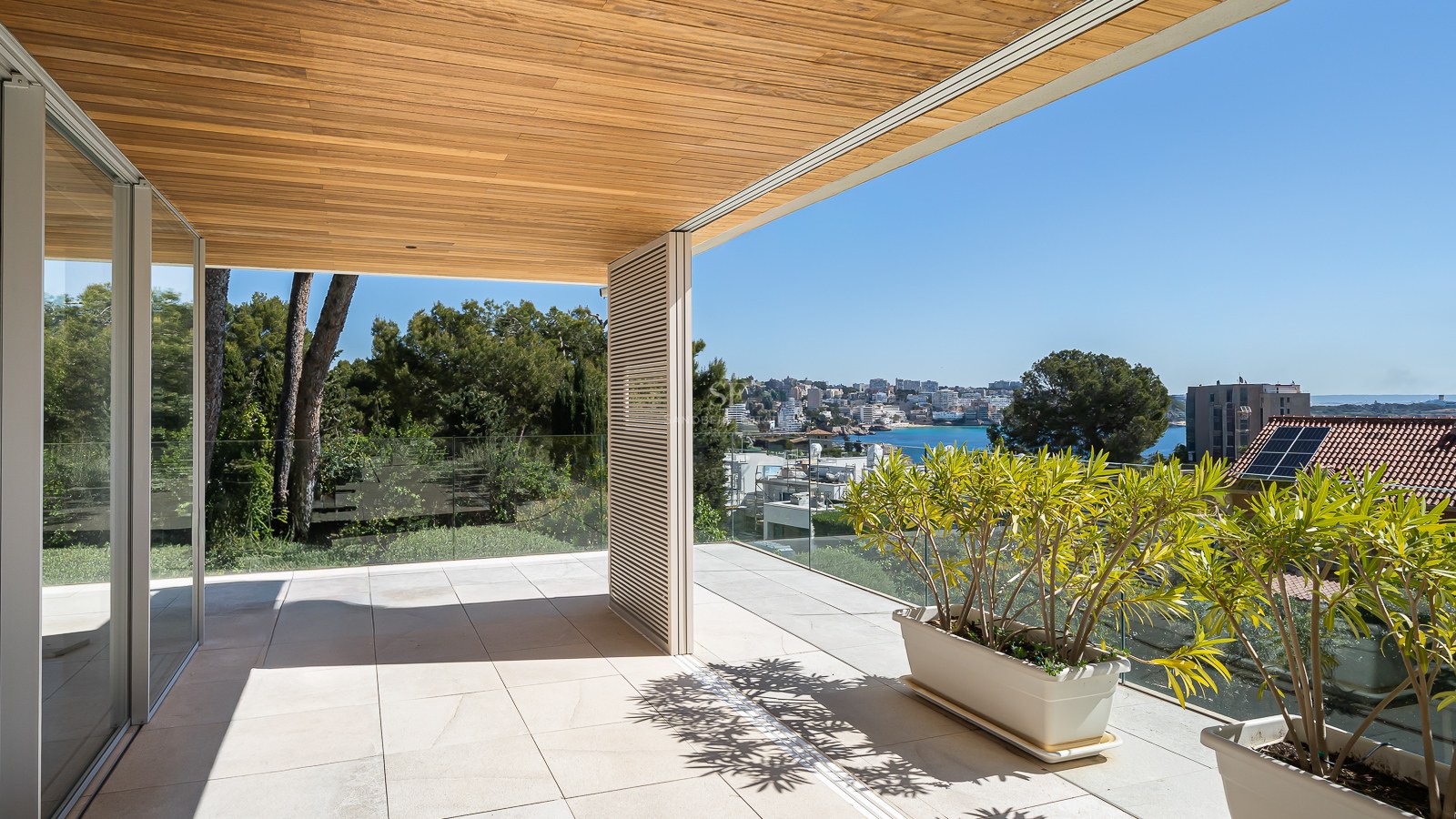 Sun-drenched modern terrace with natural stone flooring, glass railings, and a wooden ceiling overlooking a coastal town.