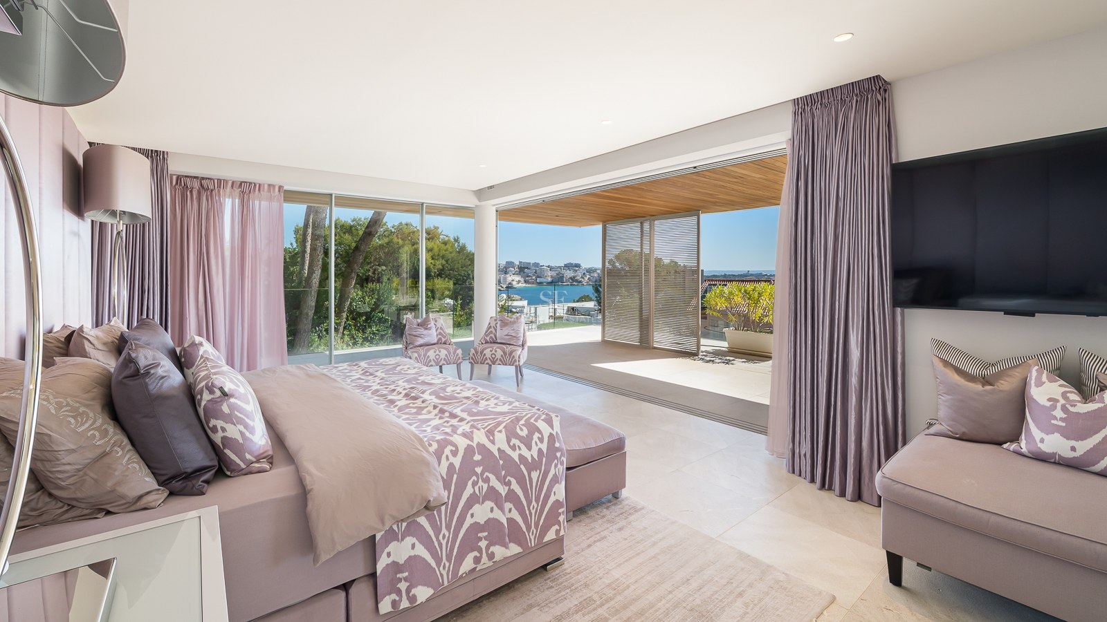 Modern bedroom with lavender accents and king bed, opening onto a sun-drenched terrace with Mediterranean coastal views.