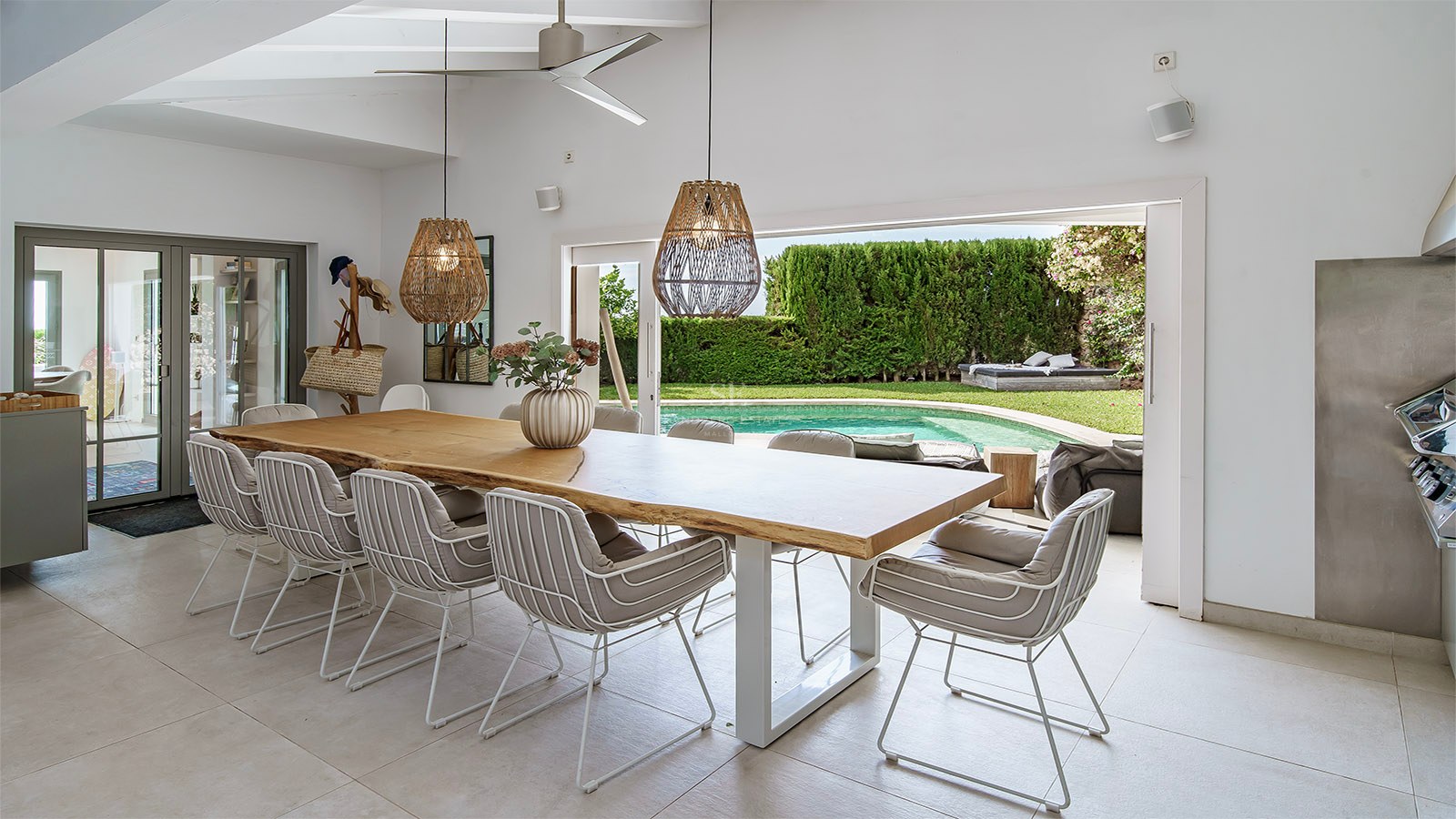 Elegante comedor con gran mesa de madera y lámparas de mimbre, abierto a una terraza con piscina y jardín.