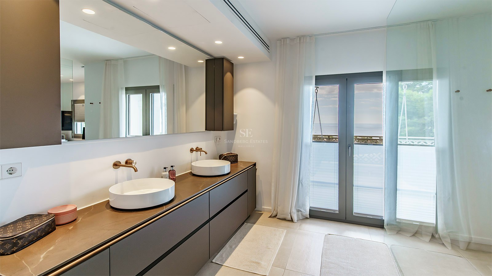Baño principal moderno con lavabo doble, gran espejo y acceso al balcón con vistas al mar.