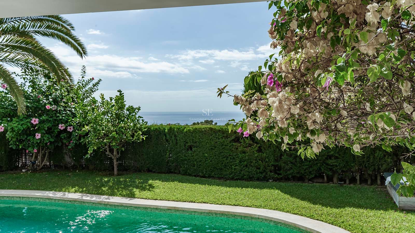 Piscina turquesa rodeada de césped verde y flores con vistas panorámicas al mar Mediterráneo.