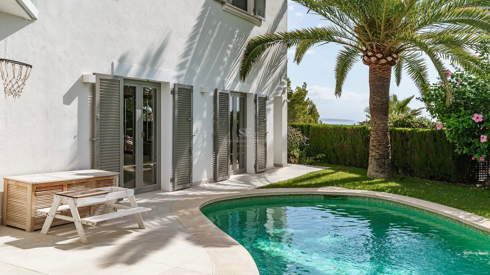 Piscina turquesa junto a una villa blanca con una palmera, persianas grises y vistas lejanas al mar.