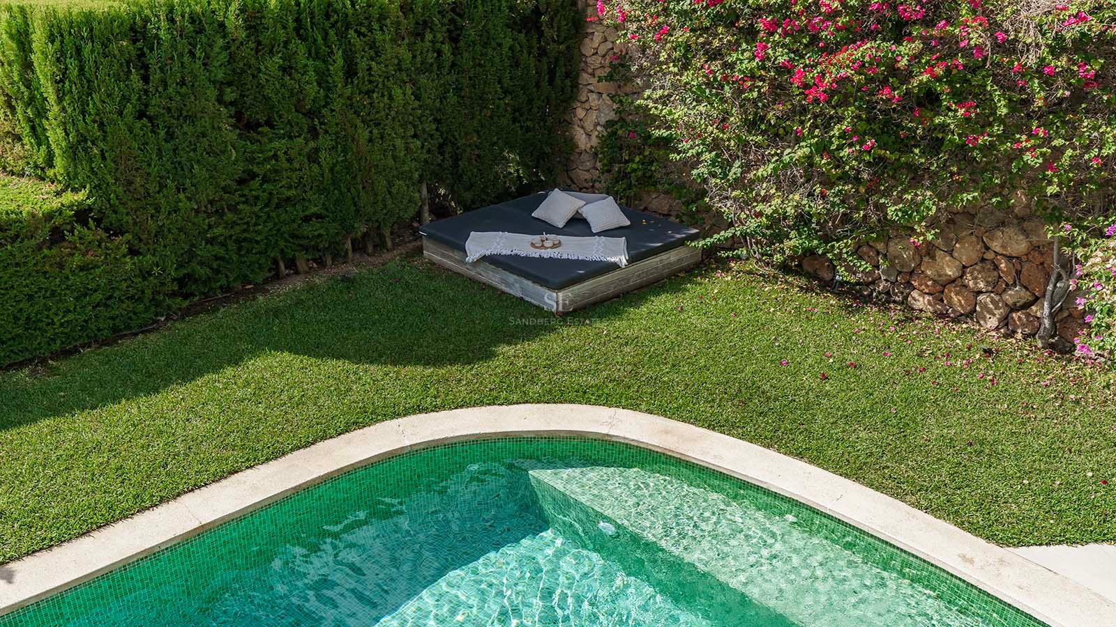 Piscina de azulejos verdes junto a un césped cuidado y una cama balinesa bajo flores de buganvilla.
