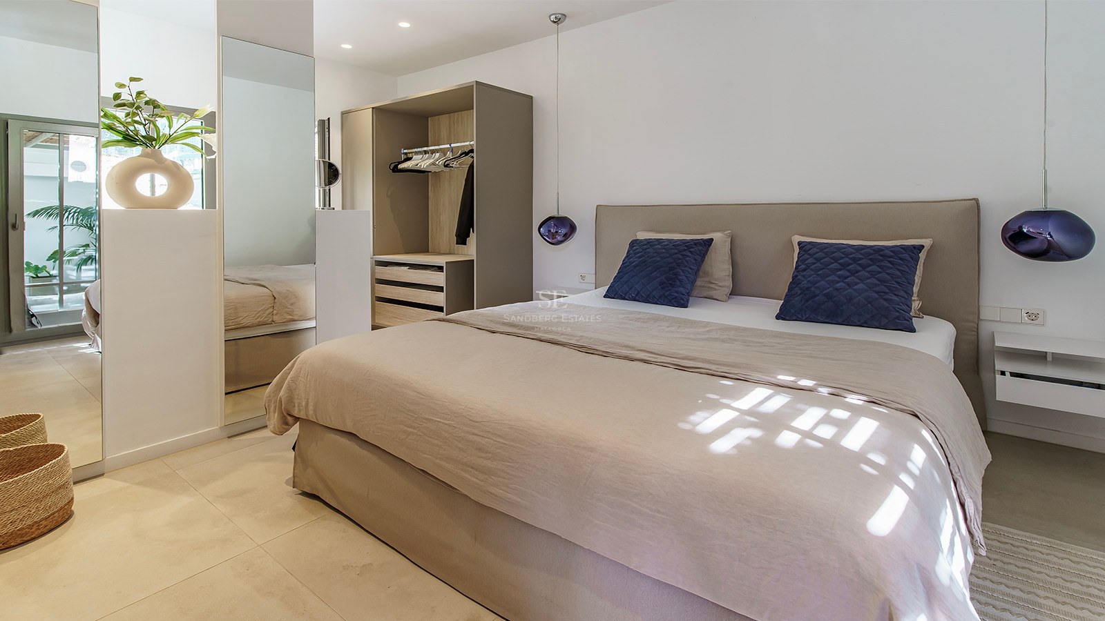 Habitación moderna y luminosa con cama beige, cojines azules, lámparas colgantes de cristal morado y espejos grandes.