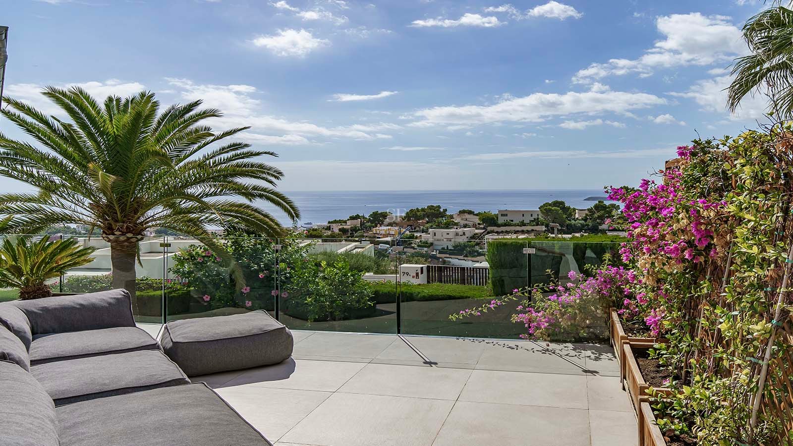 Terraza de lujo con sofá gris, barandilla de cristal, palmeras y vistas impresionantes al mar y al pueblo.