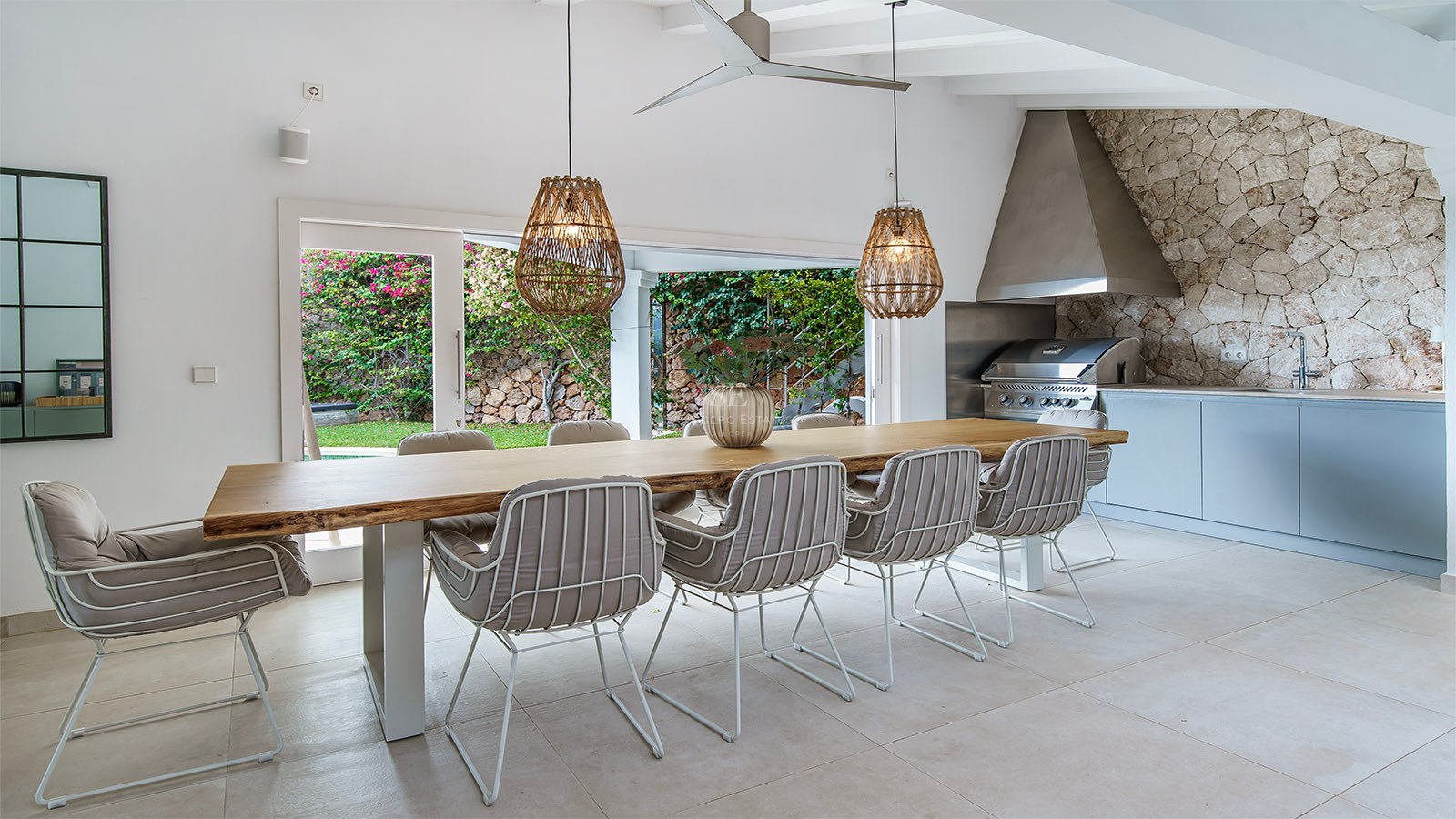 Mesa de madera maciza con sillas modernas y lámparas de mimbre junto a una cocina exterior con barbacoa de acero.