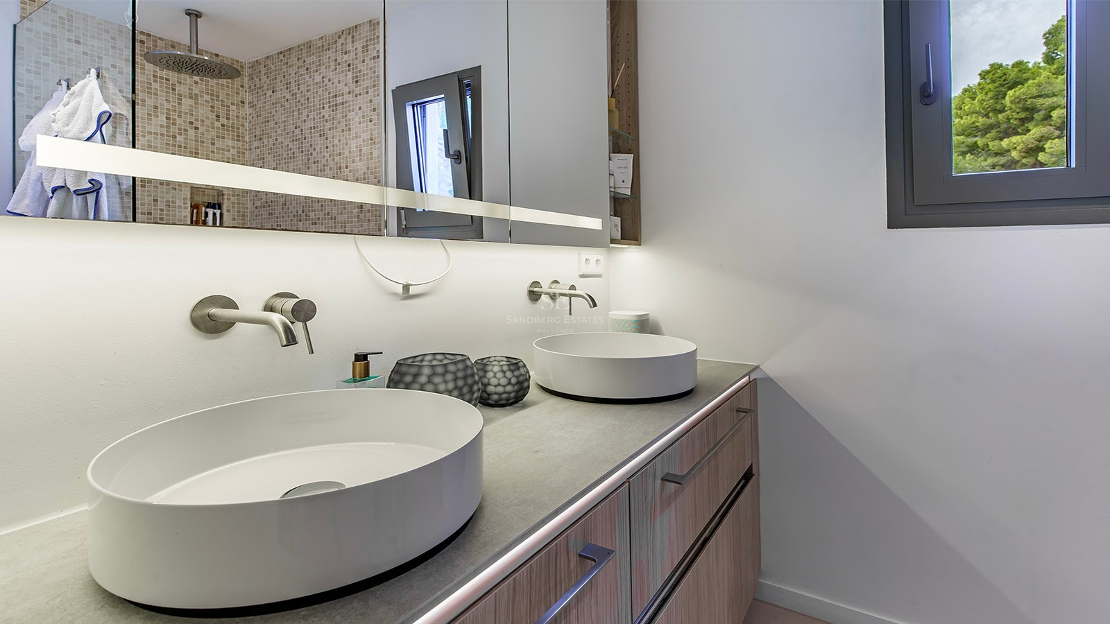 Baño moderno con dos lavabos blancos sobre encimera gris, muebles de madera y gran espejo iluminado.