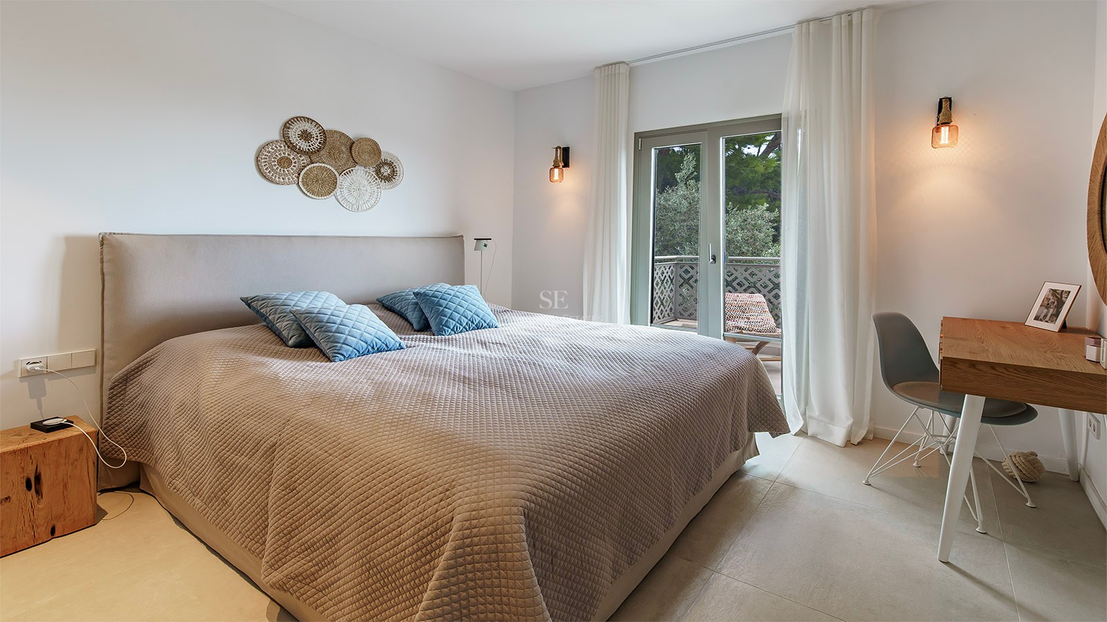 Dormitorio moderno con cama grande, colcha beige, cojines azules, escritorio de madera y puerta de cristal al balcón.