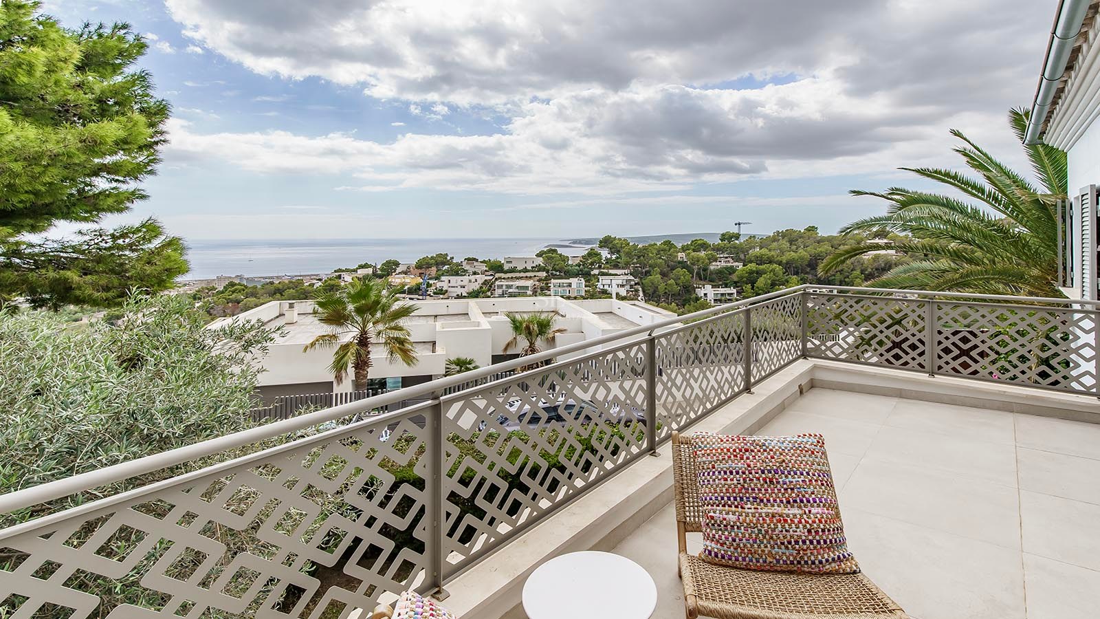 Balcón moderno con barandilla decorativa, silla de mimbre y vistas a la costa mediterránea bajo un cielo nublado.