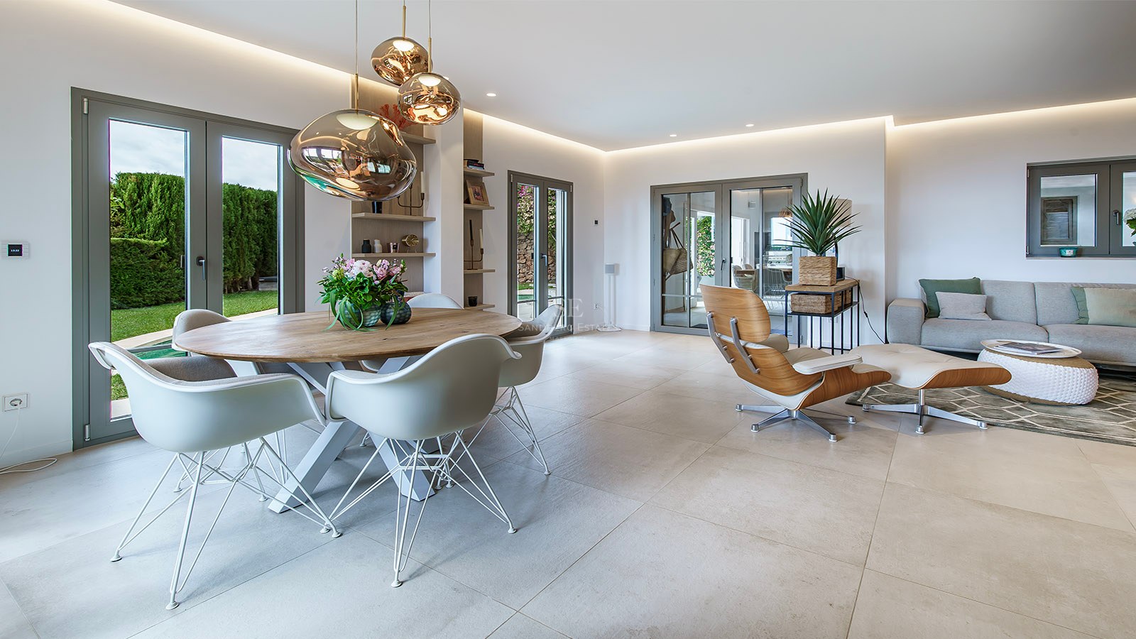 Salón moderno con comedor, sillas de diseño, lámparas de cobre y sofá gris con acceso al jardín.