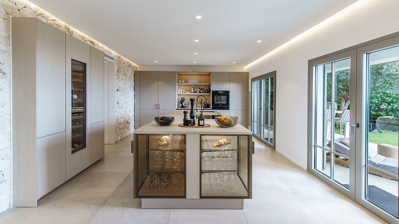 Cocina contemporánea con isla de madera, vinoteca integrada, pared de piedra natural y puertas de cristal al jardín.
