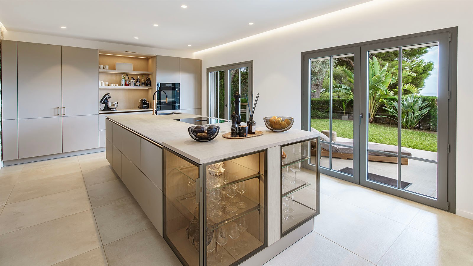 Cocina moderna con isla grande, vitrinas integradas y puertas de cristal que dan a un jardín verde.