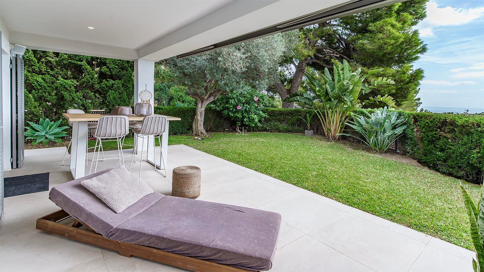 Terraza cubierta con mesa de comedor, tumbona y jardín impecable con vistas parciales al mar.