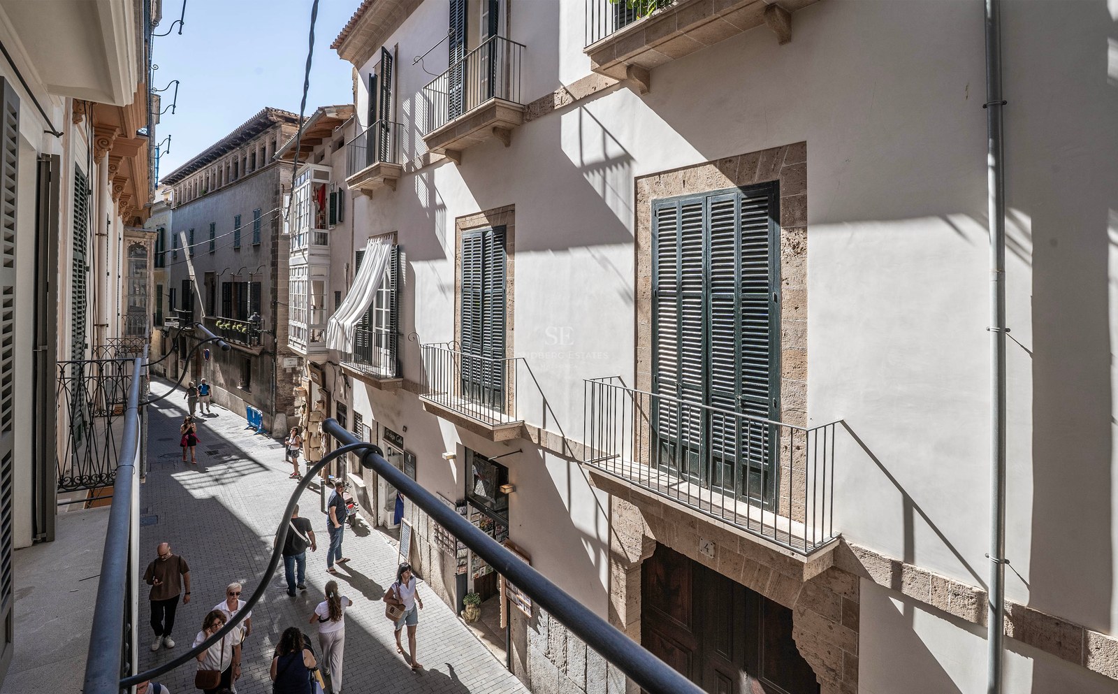 Blick von einem Balkon in eine schmale Gasse mit hellen Steingebäuden, grünen Fensterläden und Passanten.