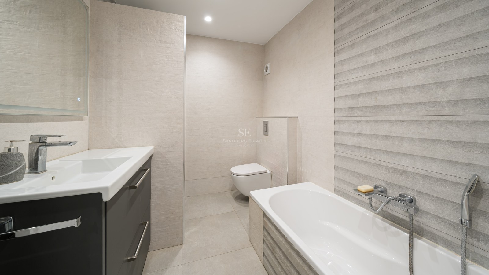 Baño moderno con bañera blanca, inodoro suspendido y tocador gris con azulejos beige texturizados.