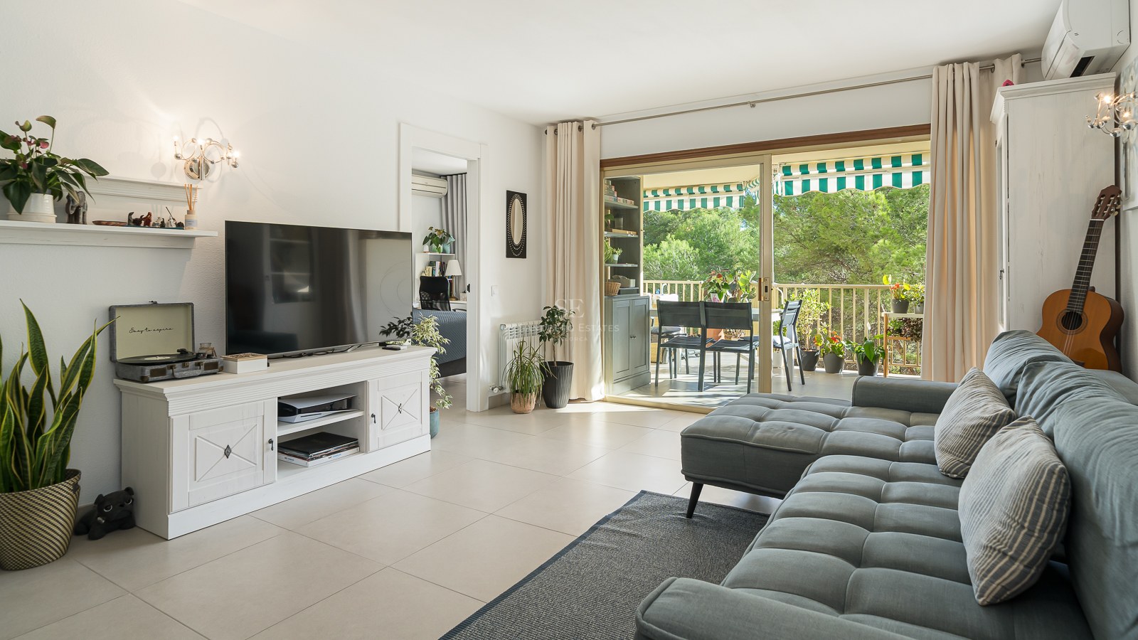 Salon moderne avec canapé gris, meuble TV blanc et baies vitrées ouvrant sur une terrasse ensoleillée.