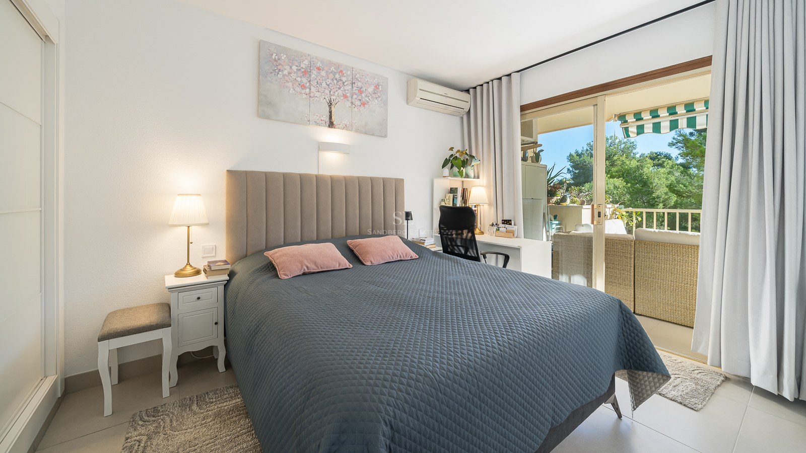 Dormitorio luminoso con cama acolchada gris, escritorio y puertas correderas de cristal que dan a un balcón soleado.
