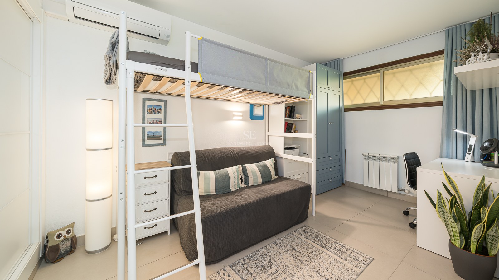Cama alta de metal blanco sobre un sofá gris, junto a un escritorio y un armario azul en una habitación luminosa.