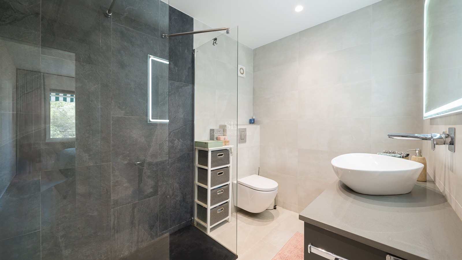 Baño moderno con ducha de cristal, azulejos gris oscuro, lavabo sobre encimera blanco y sanitario suspendido.