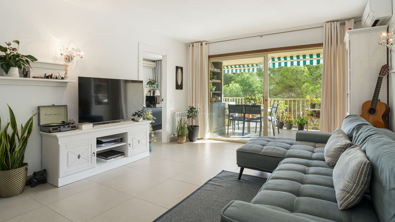 Salón amplio con sofá gris, televisión sobre mueble blanco y puertas correderas de cristal que dan a una terraza verde.