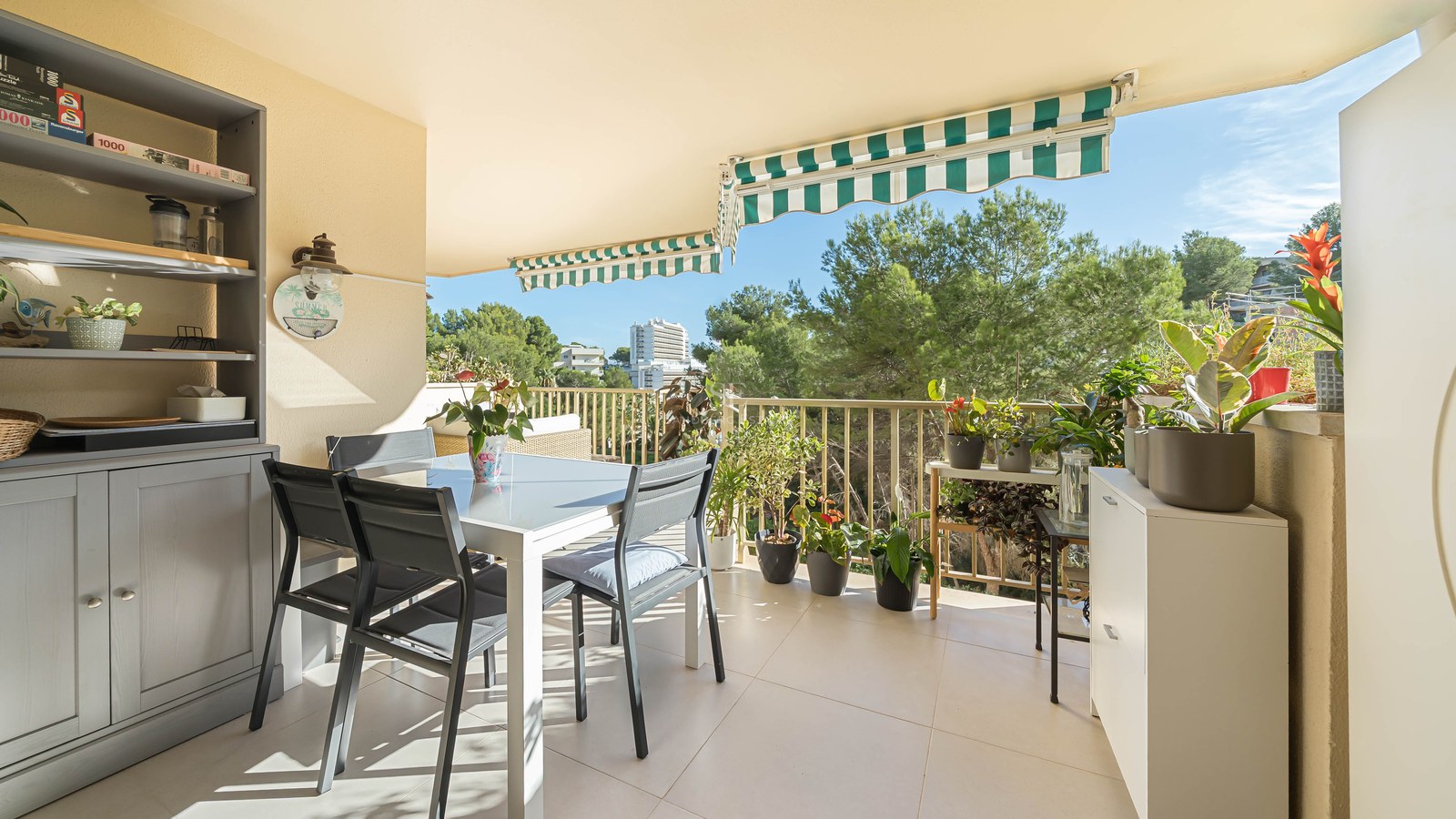 Un balcón soleado con una mesa de comedor blanca, sillas oscuras, muchas plantas en macetas y toldos de rayas verdes.